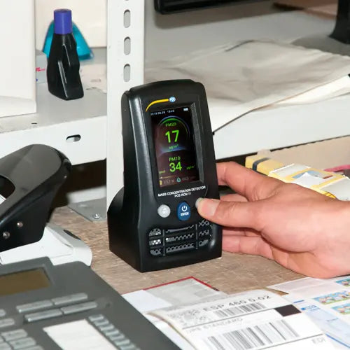Hand holding a digital air quality monitor on a desk with office equipment in the background