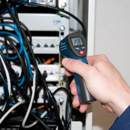 Hand holding a digital thermometer in front of electrical panels with wires.