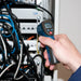 Hand holding a digital thermometer in front of electrical panels with wires.