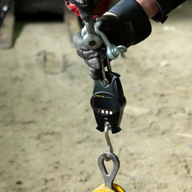 Digital weight scale with a hook attached to a person's gloved hand on a blurred background