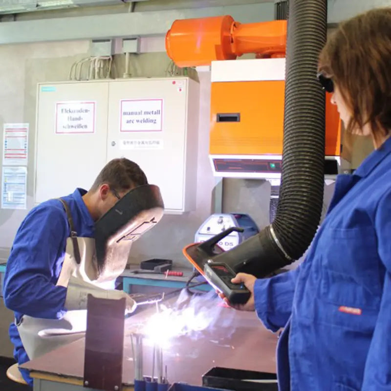 Two workers in a manufacturing setting with one using a welding torch and the other operating a machine.