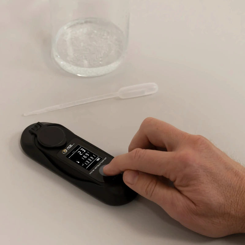 Hand using a refractometer on a white surface with a glass container and straw in the background.