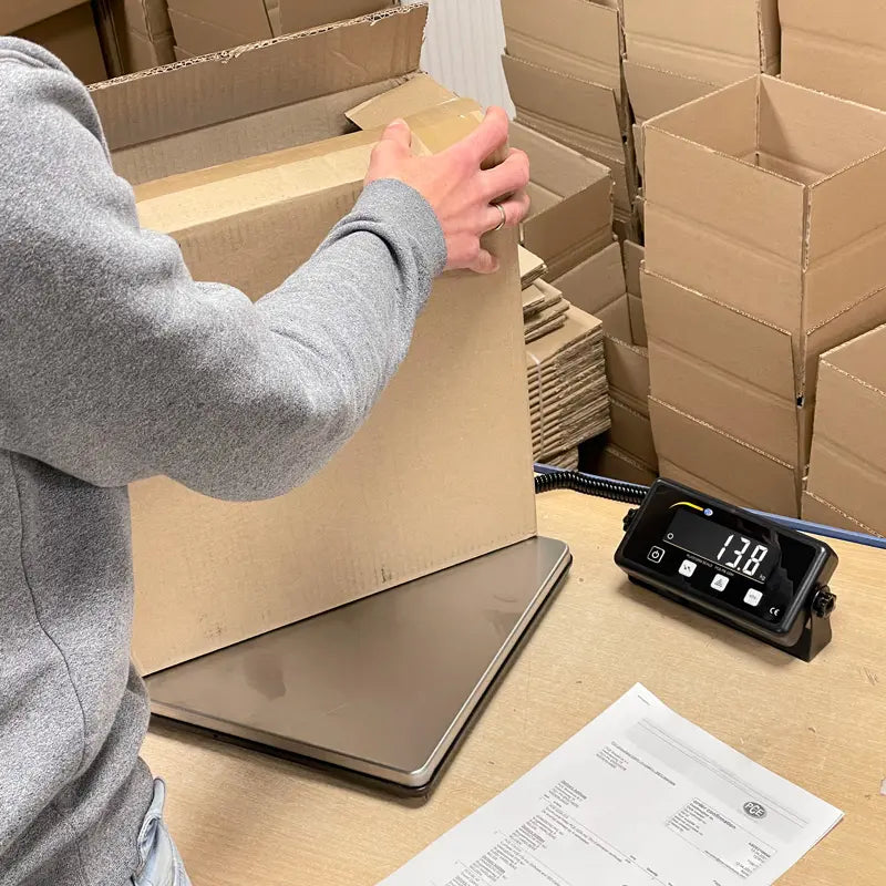 Person packaging a box on a digital scale with stacked boxes in the background