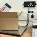 Cardboard box being weighed on a digital scale with a power outlet in the background.